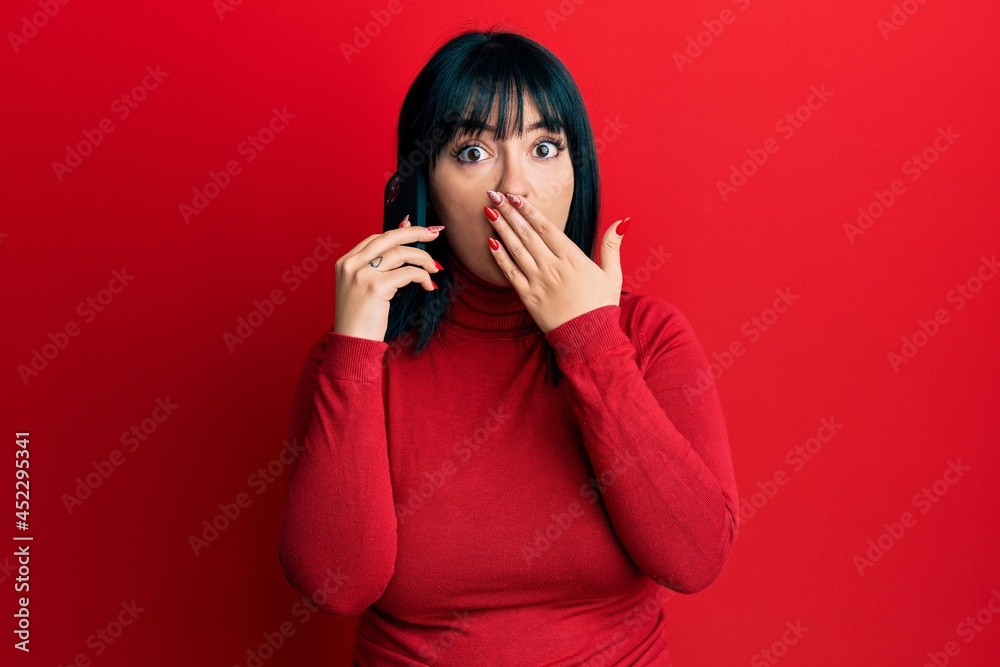 Fototapeta premium Young hispanic woman having conversation talking on the smartphone covering mouth with hand, shocked and afraid for mistake. surprised expression