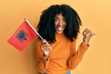 African american woman with afro hair holding albania flag pointing thumb up to the side smiling happy with open mouth