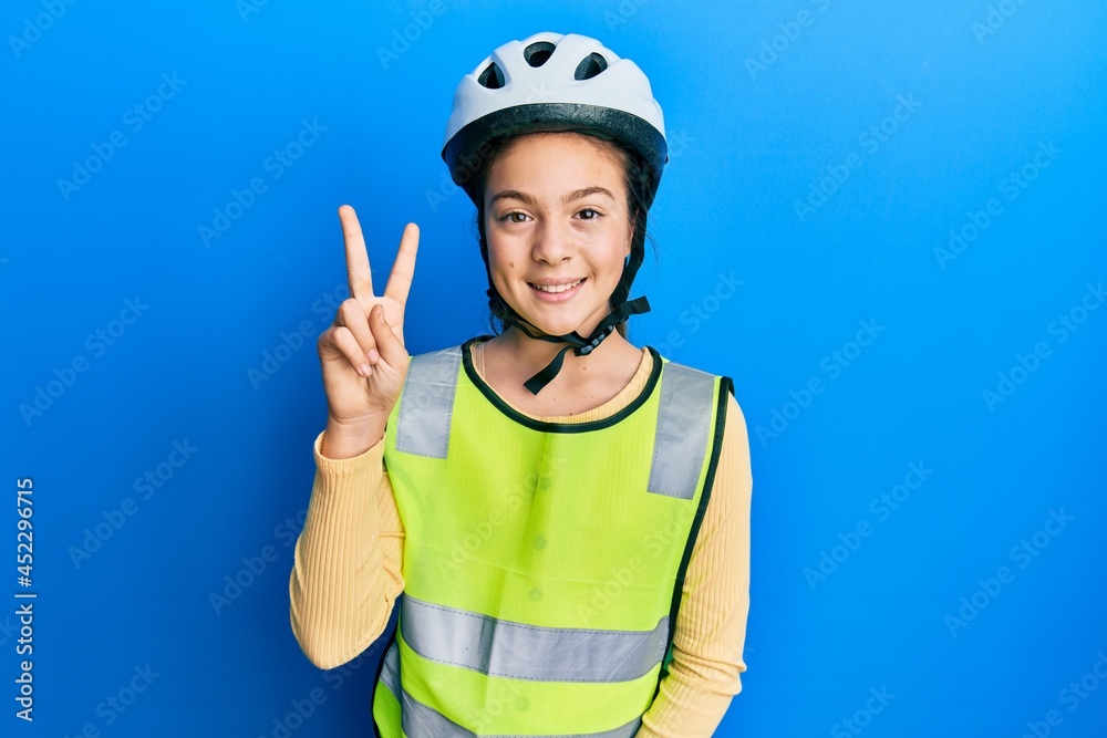 Beautiful brunette little girl wearing bike helmet and reflective vest showing and pointing up with fingers number two while smiling confident and happy. Beautiful brunette little girl wearing bike helmet and reflective vest showing and pointing up with fingers number two while smiling confident and happy.