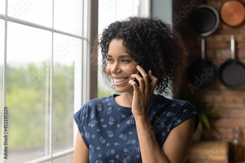 Happy thoughtful millennial Black mixed race woman making telephone call, looking out of window, thinking over mobile phone talk, enjoying conversation with toothy smile, chatting to friend,