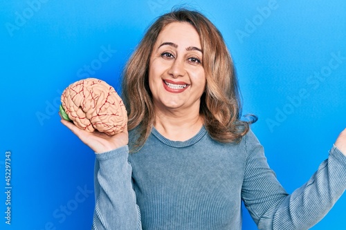 Middle age caucasian woman holding brain celebrating achievement with happy smile and winner expression with raised hand