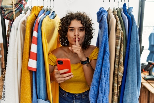 Young hispanic woman searching clothes on clothing rack using smartphone asking to be quiet with finger on lips. silence and secret concept.