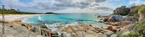 Panorama of beach with turquoise water, cloudy blue sky, orange rocky shoreline. Bay of Fires, Tasmania, Australia. No people