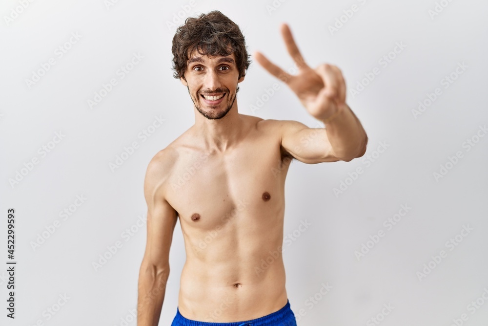 Young hispanic man standing shirtless over isolated, background smiling looking to the camera showing fingers doing victory sign. number two.