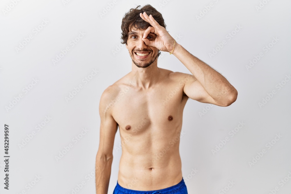 Fototapeta premium Young hispanic man standing shirtless over isolated, background doing ok gesture with hand smiling, eye looking through fingers with happy face.