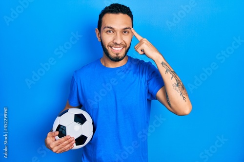 Hispanic man with beard holding soccer ball smiling pointing to head with one finger, great idea or thought, good memory