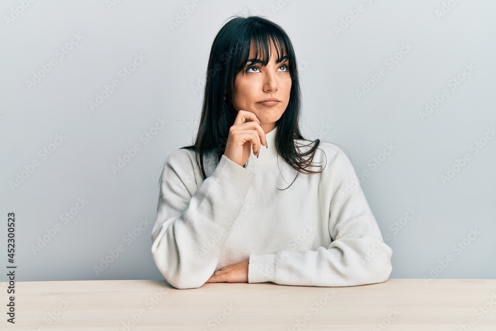 Young brunette woman with bangs wearing casual clothes sitting on the table thinking concentrated about doubt with finger on chin and looking up wondering Young brunette woman with bangs wearing casual clothes sitting on the table thinking concentrated about doubt with finger on chin and looking up wondering