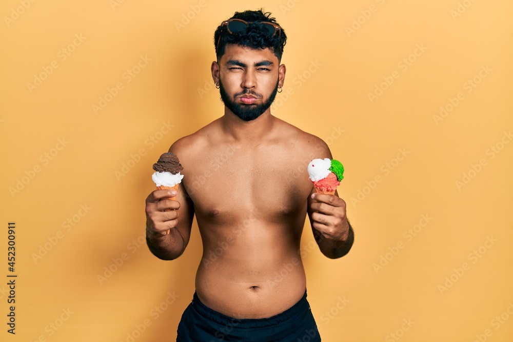 Arab man with beard wearing swimwear eating two ice cream cones puffing cheeks with funny face. mouth inflated with air, catching air. Arab man with beard wearing swimwear eating two ice cream cones puffing cheeks with funny face. mouth inflated with air, catching air.