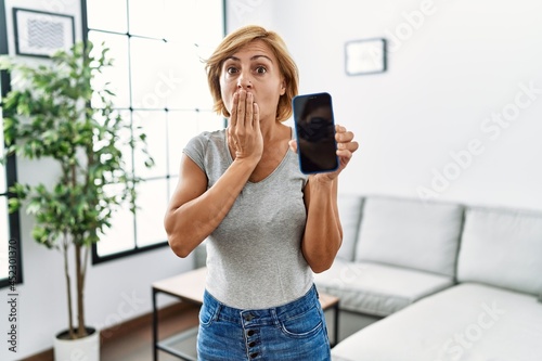 Middle age blonde woman working holding smartphone showing screen covering mouth with hand, shocked and afraid for mistake. surprised expression