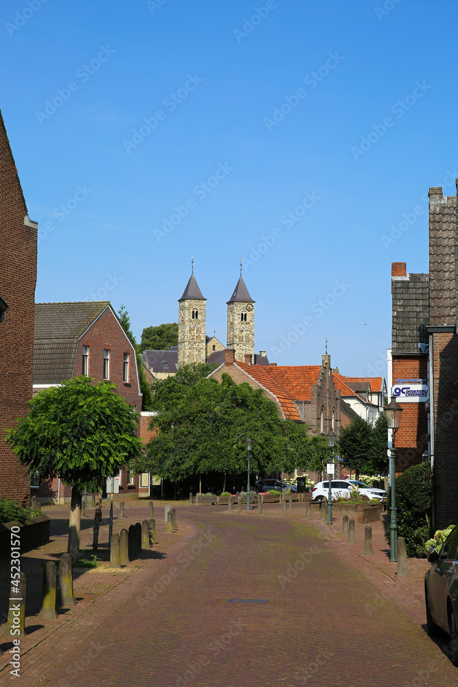 Fototapeta premium Sint Odilienberg, Netherlands - 9. July 2021: View over street beyond houses on roman basilica from 11th century against blue summer sky