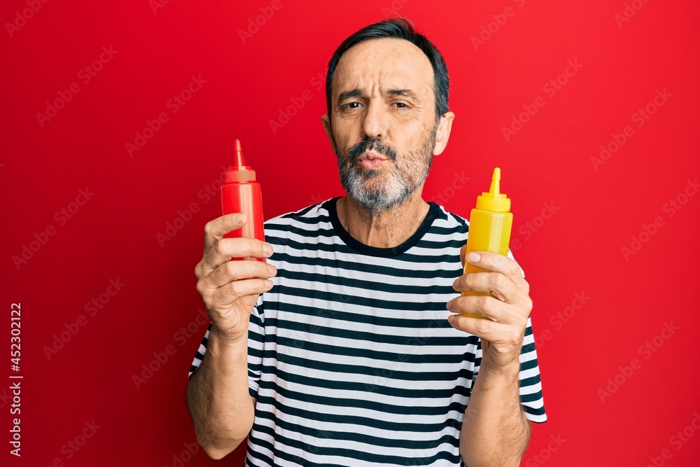 Middle age hispanic man holding ketchup and mustard bottle looking at the camera blowing a kiss being lovely and sexy. love expression.