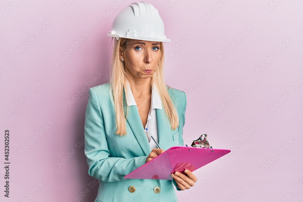 Young blonde woman wearing architect hardhat writing on clipboard depressed and worry for distress, crying angry and afraid. sad expression. Young blonde woman wearing architect hardhat writing on clipboard depressed and worry for distress, crying angry and afraid. sad expression.