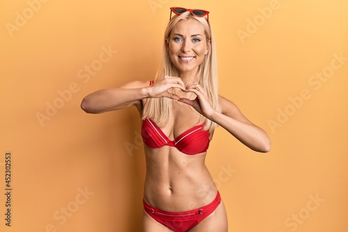 Young blonde woman wearing bikini smiling in love doing heart symbol shape with hands. romantic concept.