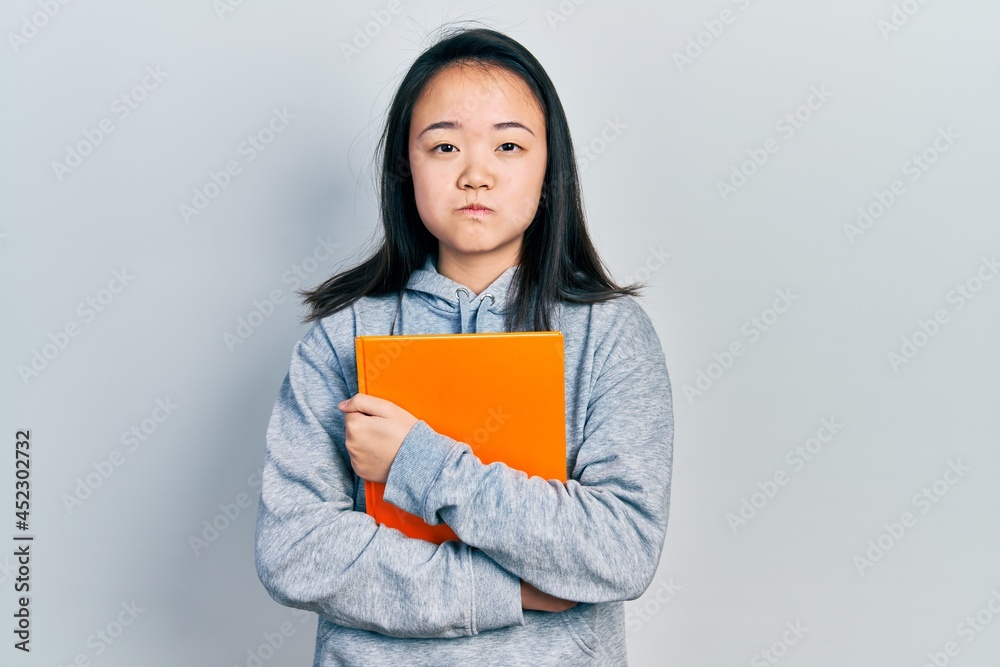 Young chinese girl holding book puffing cheeks with funny face. mouth inflated with air, catching air. Young chinese girl holding book puffing cheeks with funny face. mouth inflated with air, catching air.