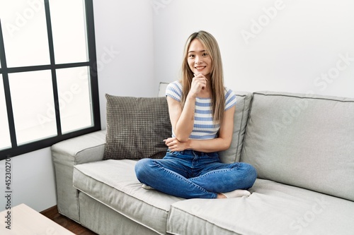 Asian young woman sitting on the sofa at home looking confident at the camera smiling with crossed arms and hand raised on chin. thinking positive.