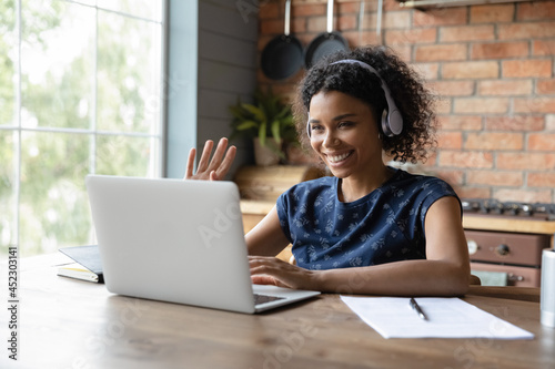 Happy Black student girl in wireless earphones attending virtual webinar, learning conference, waving hello at laptop webcam. Coach, teacher blogger giving online training, greeting audience