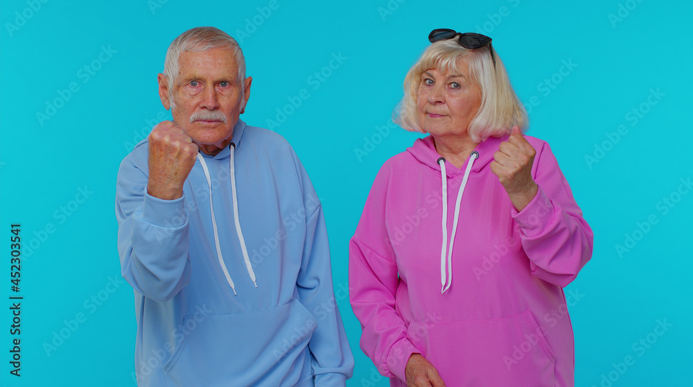 Aggressive angry senior woman granny with man grandfather trying to fight at camera, shaking fist, boxing with expression, punishment isolated on blue background. Elderly grandmother with grandpa Aggressive angry senior woman granny with man grandfather trying to fight at camera, shaking fist, boxing with expression, punishment isolated on blue background. Elderly grandmother with grandpa