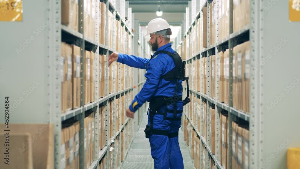 Warehouse worker in ergoskeleton is storaging a box into the shelf Warehouse worker in ergoskeleton is storaging a box into the shelf