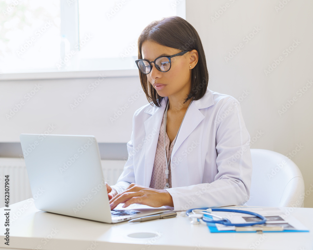 Focused african american female doctor in eyeglasses using laptop while sitting at workplace in medical clinic, young afro woman physician dressed in white coat working on computer in hospital Focused african american female doctor in eyeglasses using laptop while sitting at workplace in medical clinic, young afro woman physician dressed in white coat working on computer in hospital