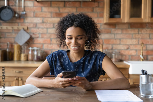 Happy Afro American student girl using smartphone at kitchen table, reading, texting, looking at screen. Black young woman, remote employee chatting online, checking messages, working from home