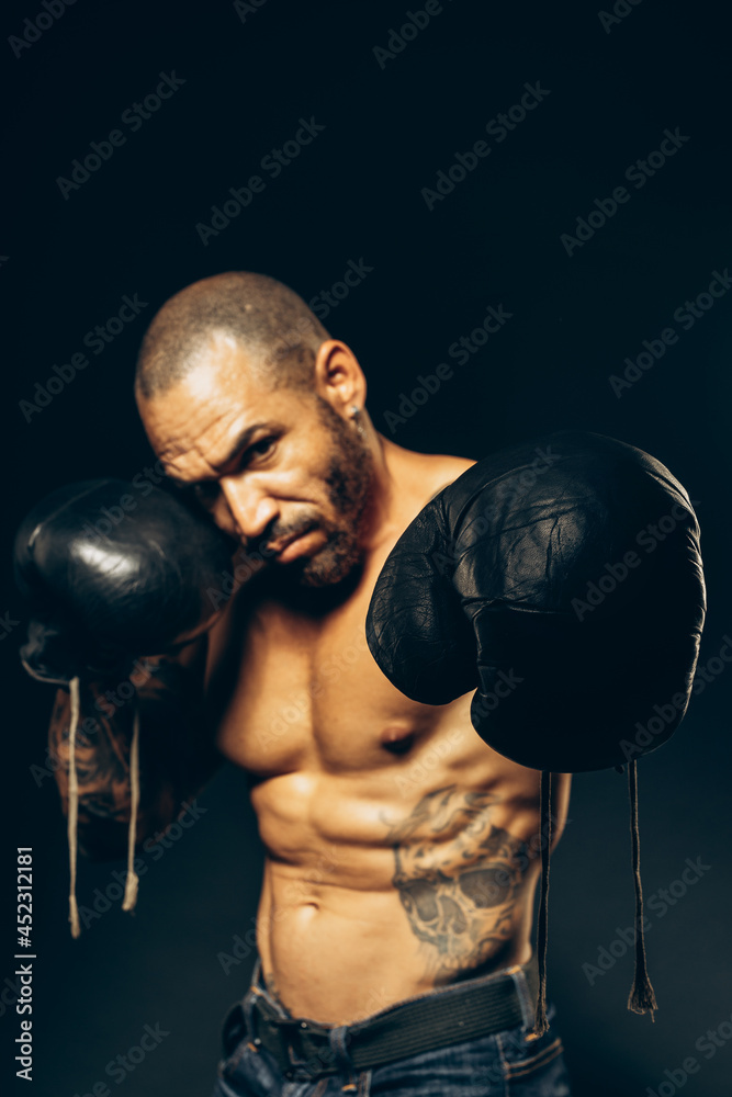 Studio portrait of a muscular boxer with a naked torso on a dark background. The Latin American puncher's attack. A serious fighter performs punches.