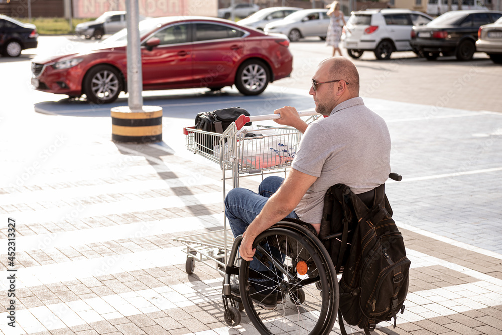 Obraz premium Adult disabled man in a wheelchair pushes a cart towards a car in a supermarket parking lot