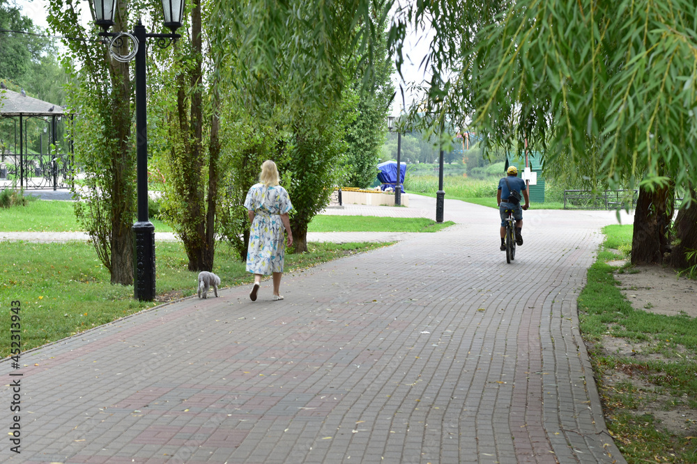 A cyclist is riding along the path in the park and a slender woman is walking.