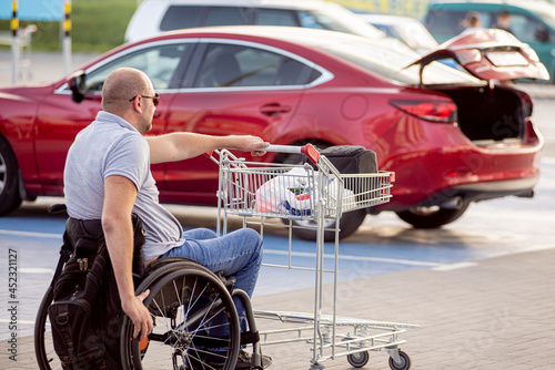 Adult disabled man in a wheelchair pushes a cart towards a car in a supermarket parking lot