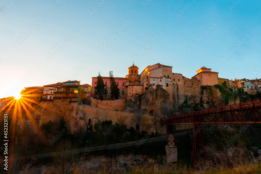 Cityscape in old town changing light at sunset; Hanging houses with old town hanging over mountain and red bridge of San Pablo de Cuenca with sun star, world heritage site, Spain. Horizontal view