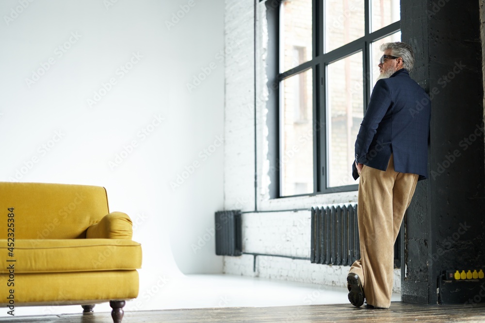 Full length shot of stylish middle aged man in business casual wear and sunglasses leaning on the wall, looking away while posing in loft interior