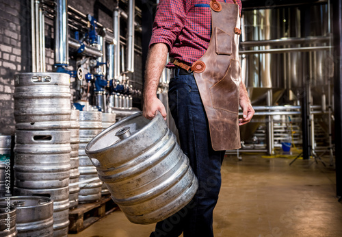 Young male brewer in leather apron holds barrel with craft beer at modern brewery factory