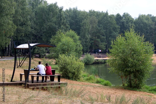 A young couple are sitting on the shore of a pond in a gazebo. Summer warm day.