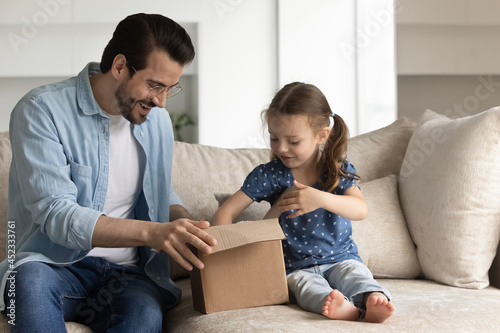 Happy dad and excited daughter girl unboxing parcel with surprising gift, opening cardboard box, looking inside container, producing goods. Father and kid receiving purchase via delivery service
