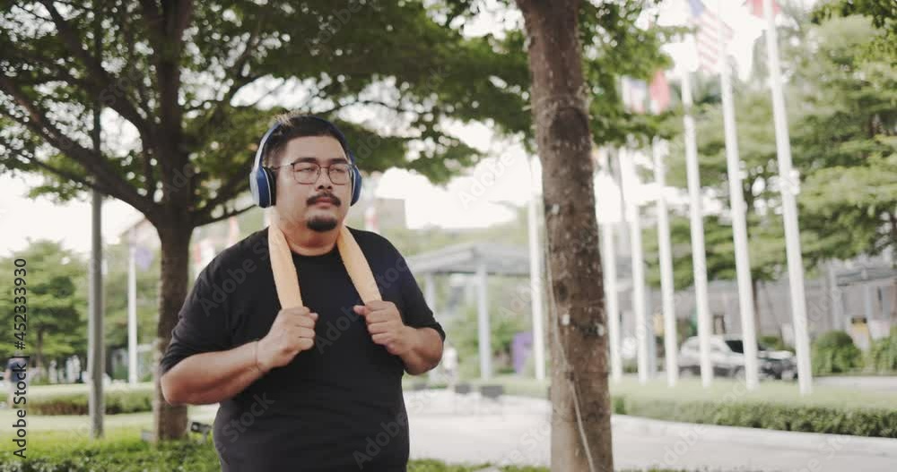 Obesity Asian man wearing active music headphone running in park morning. Jogging workout to burn body fat to healthy. Commitment to body care for good health. Weekend activity happy lifestyle concept Obesity Asian man wearing active music headphone running in park morning. Jogging workout to burn body fat to healthy. Commitment to body care for good health. Weekend activity happy lifestyle concept