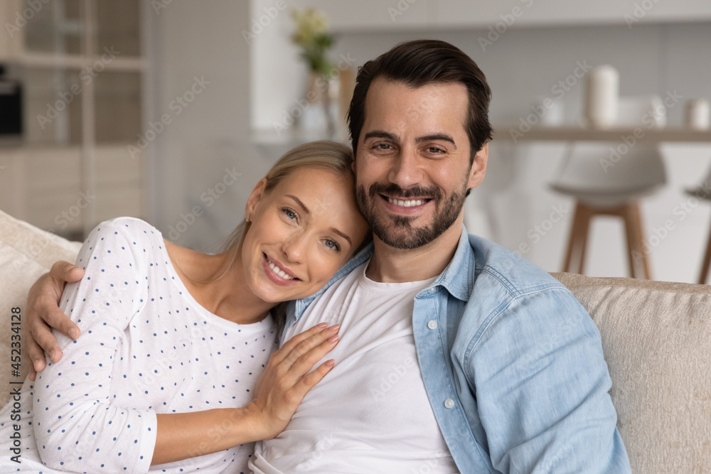 Happy millennial married couple of homeowners head shot portrait. Husband and wife sitting on couch, looking at camera with toothy smile, hugging with love, relaxing in new comfortable house Happy millennial married couple of homeowners head shot portrait. Husband and wife sitting on couch, looking at camera with toothy smile, hugging with love, relaxing in new comfortable house