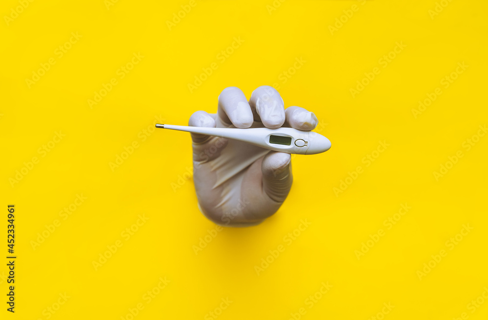 A hand in a white medical glove is pushed through a torn hole in yellow paper and holds an electronic thermometer. The concept of diagnosing colds, covid-19, coronavirus. Copy space. A hand in a white medical glove is pushed through a torn hole in yellow paper and holds an electronic thermometer. The concept of diagnosing colds, covid-19, coronavirus. Copy space.