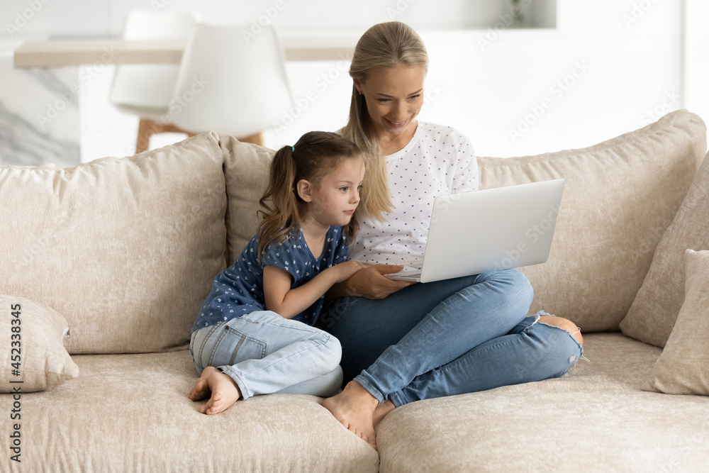 Happy young blonde mom and gen Z daughter kid using laptop together, virtual app on computer, resting on sofa in cozy living room, making video call, watching movie, shopping online, playing game Happy young blonde mom and gen Z daughter kid using laptop together, virtual app on computer, resting on sofa in cozy living room, making video call, watching movie, shopping online, playing game
