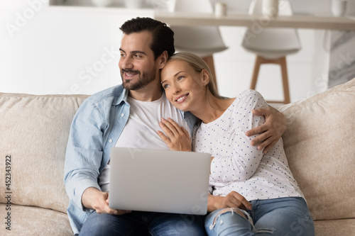 Happy couple thinking of family future, dreaming of home owning, planning mortgage. Millennial husband and wife relaxing on couch, using laptop, holding computer, looking away, smiling