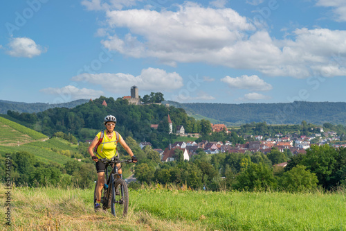 happy senior woman riding her electric mountain bike on sunny day in the Bottwartal Valley with beautiful medieval castle in the background, Beilstein near Heilbronn, Baden-Wuerttemberg, Germany