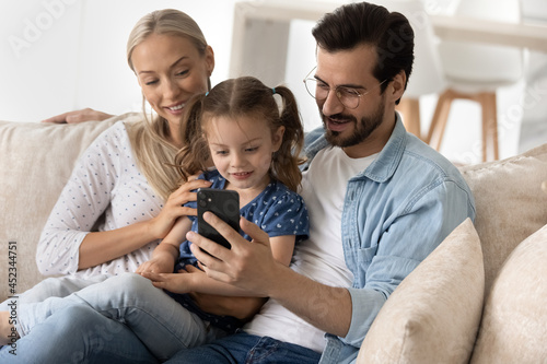 Happy parents and sweet little gen Z kid using app on smartphone together, hugging on couch at home, making online video call, watching media content. Family with digital gadget resting on sofa