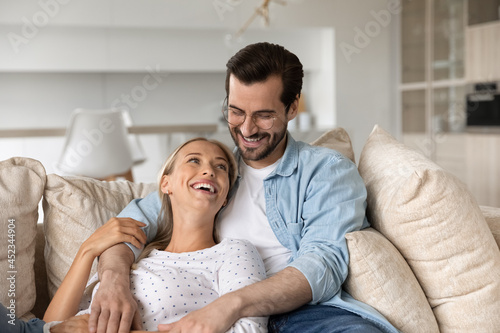 Happy millennial couple relaxing on soft comfortable couch, hugging, talking, laughing, enjoying dating, living together. Husband embracing wife. Family relationships, romance at home concept