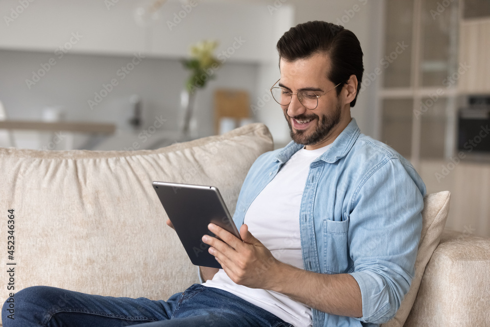 Happy user holding digital device, resting on couch, relaxing at home. Relaxed millennial man using tablet computer, making video call, reading book online, watching virtual content, smiling at screen Happy user holding digital device, resting on couch, relaxing at home. Relaxed millennial man using tablet computer, making video call, reading book online, watching virtual content, smiling at screen