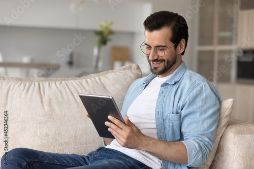 Happy user holding digital device, resting on couch, relaxing at home. Relaxed millennial man using tablet computer, making video call, reading book online, watching virtual content, smiling at screen