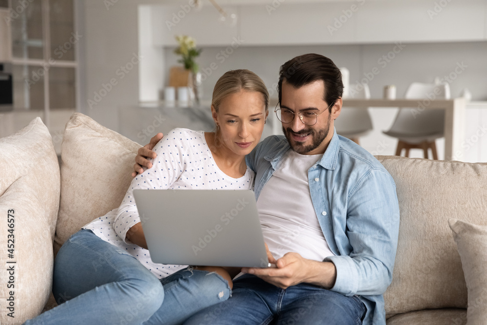 Happy millennial couple focused on laptop sitting together on sofa at home, hugging, using app on computer, watching movie online, shopping on internet, paying bills, insurance, mortgage fee Happy millennial couple focused on laptop sitting together on sofa at home, hugging, using app on computer, watching movie online, shopping on internet, paying bills, insurance, mortgage fee