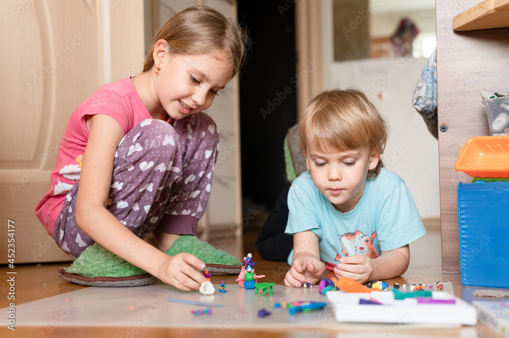 Russia, Moscow, October 2020 - two little happy kids a boy of four year old and a girl of seven year old siblings or friends at home on floor together playing and sculpting with plasticine Russia, Moscow, October 2020 - two little happy kids a boy of four year old and a girl of seven year old siblings or friends at home on floor together playing and sculpting with plasticine