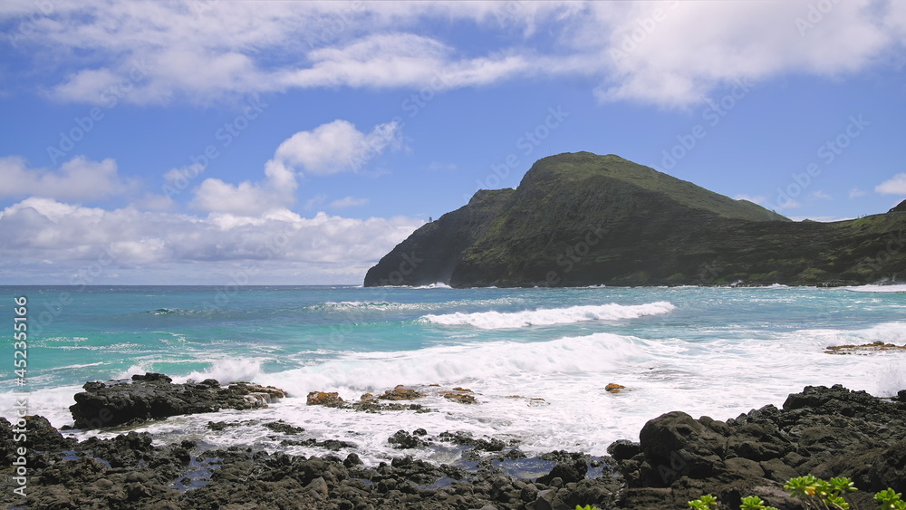 View of makapuu lighthouse. Waves of Pacific Ocean wash over yellow sand of tropical beach. Magnificent mountains of Hawaiian island of Oahu against backdrop of blue sky with white clouds.