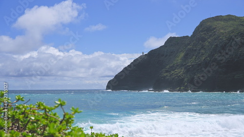 View of makapuu lighthouse. Waves of Pacific Ocean wash over yellow sand of tropical beach. Magnificent mountains of Hawaiian island of Oahu against backdrop of blue sky with white clouds.
