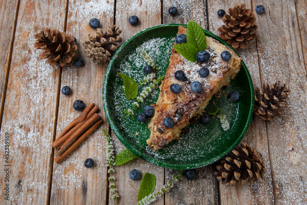 Piece of tasty cake with blueberries on wooden table with cones, cinnamon and mint. New Year Eve vintage meal Piece of tasty cake with blueberries on wooden table with cones, cinnamon and mint. New Year Eve vintage meal