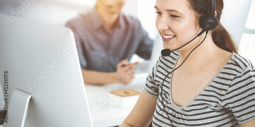 Принти на полотні Casual dressed young woman using headset and computer while talking with customers online