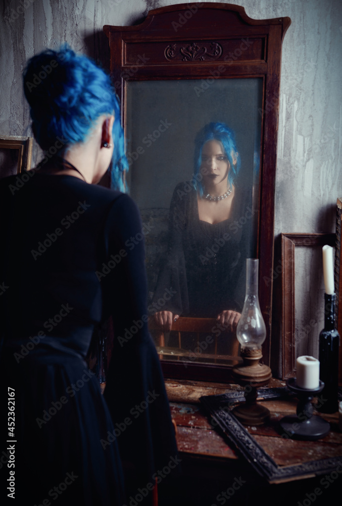 Indoors portrait of lovely goth girl in black dress. Blue-haired gothic lady looking into old dirty mirror. Vintage style